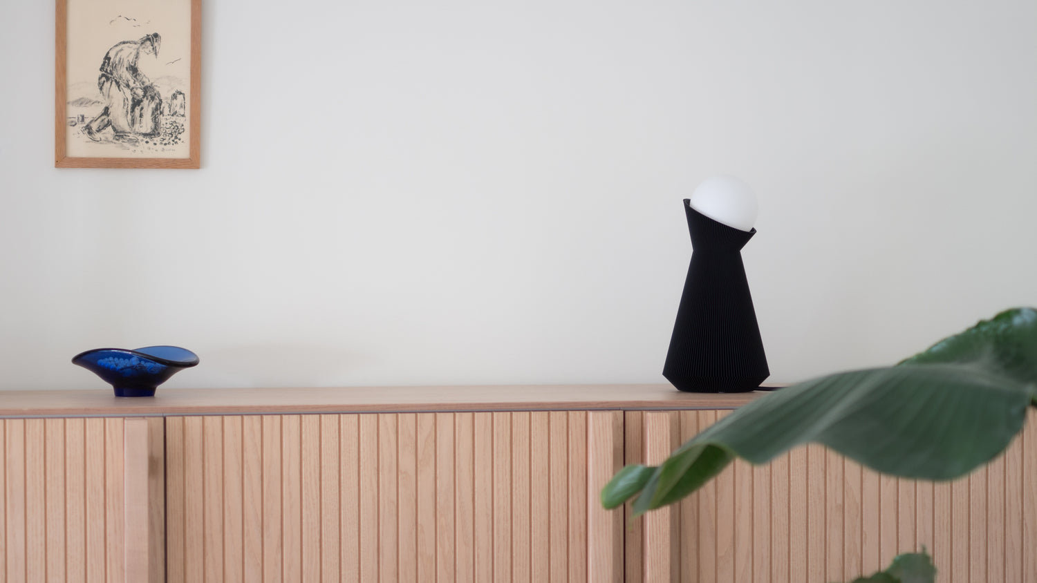Charcoal Sera lamp on a wooden shelf with a plant and framed picture in the background