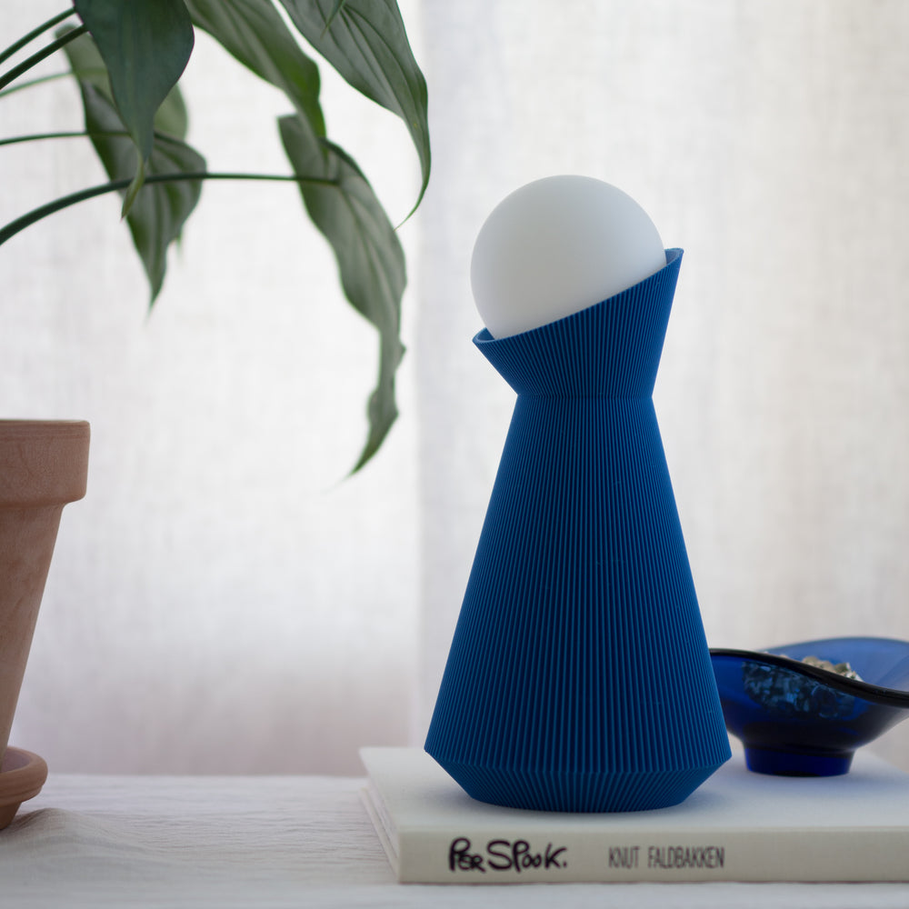 Denim Blue sera lamp with a white light bulb, placed on a surface with a plant and books in the background.